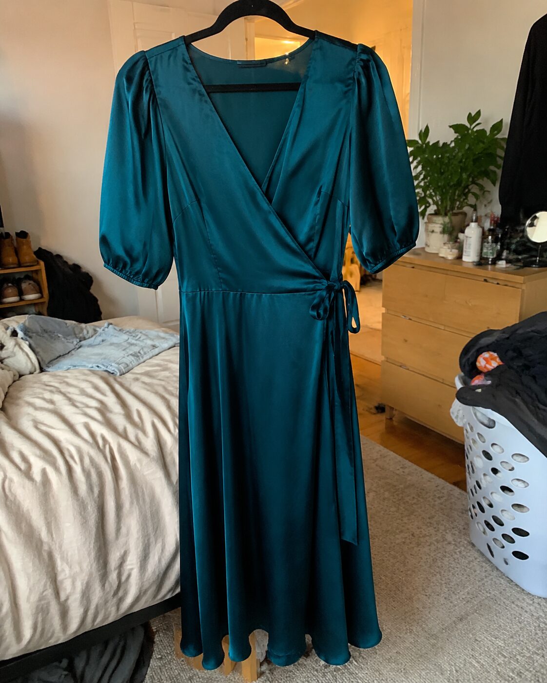 Teal satin wrap dress photographed on a hanger in a cluttered bedroom